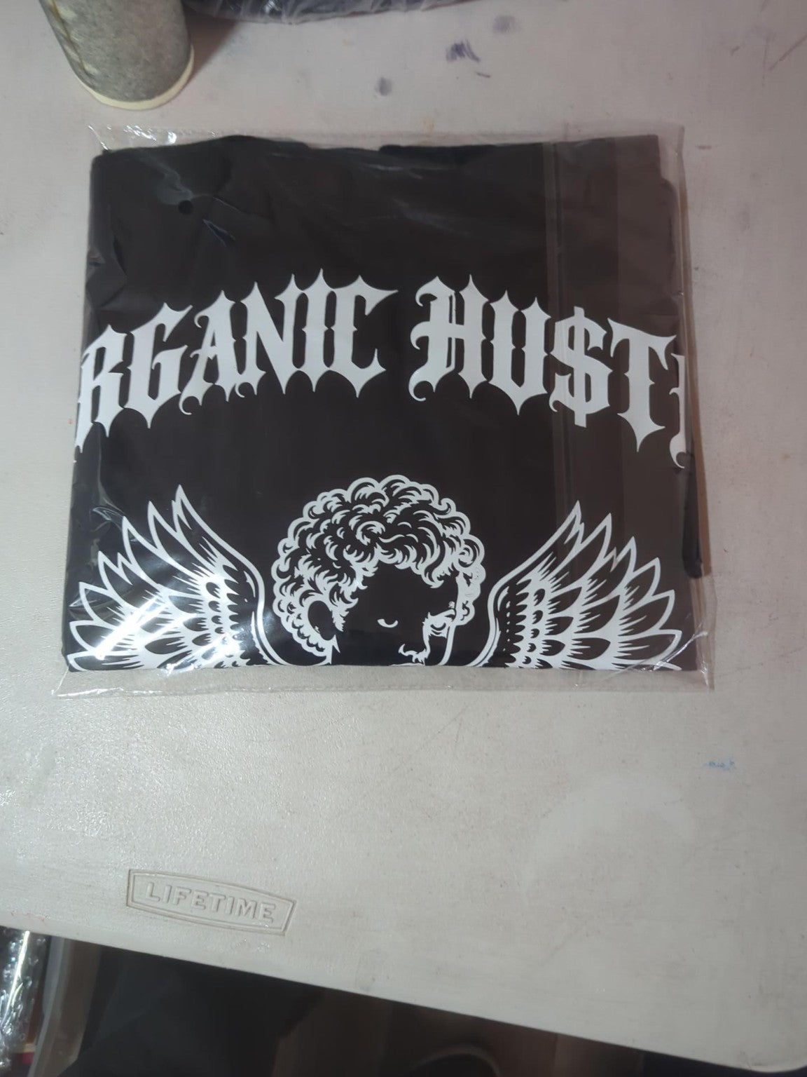 Actual packaged black Guardian Angel tee with white Gothic Organic Hu$tle lettering and winged cherub design