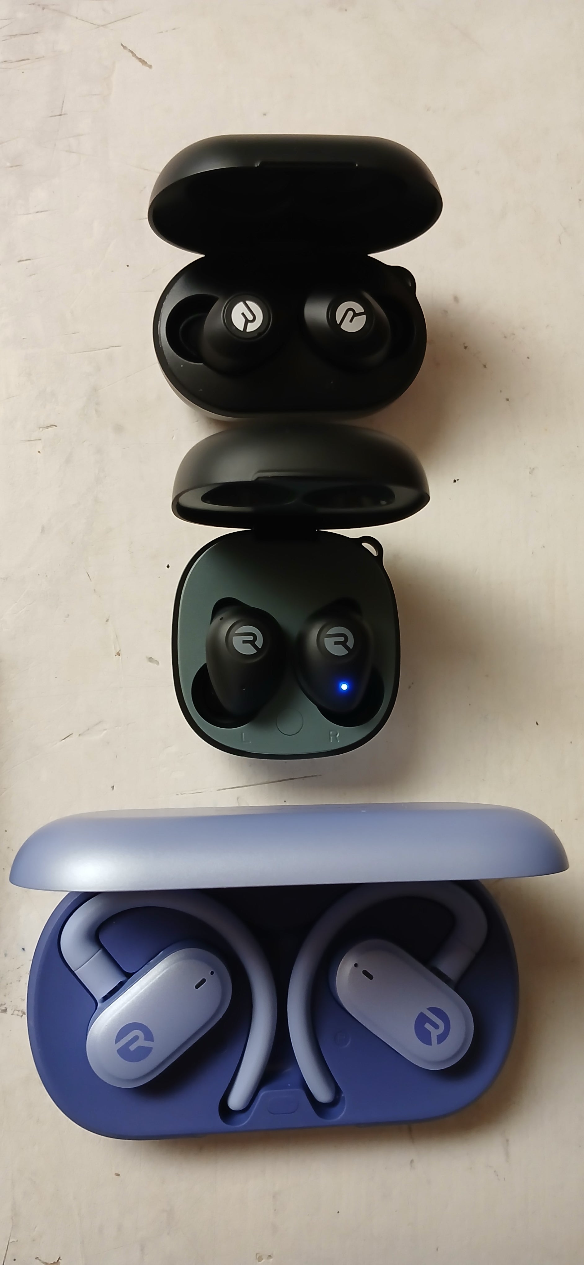 Three pairs of wireless earbuds, two black pairs and one with a blue case, on a light surface.