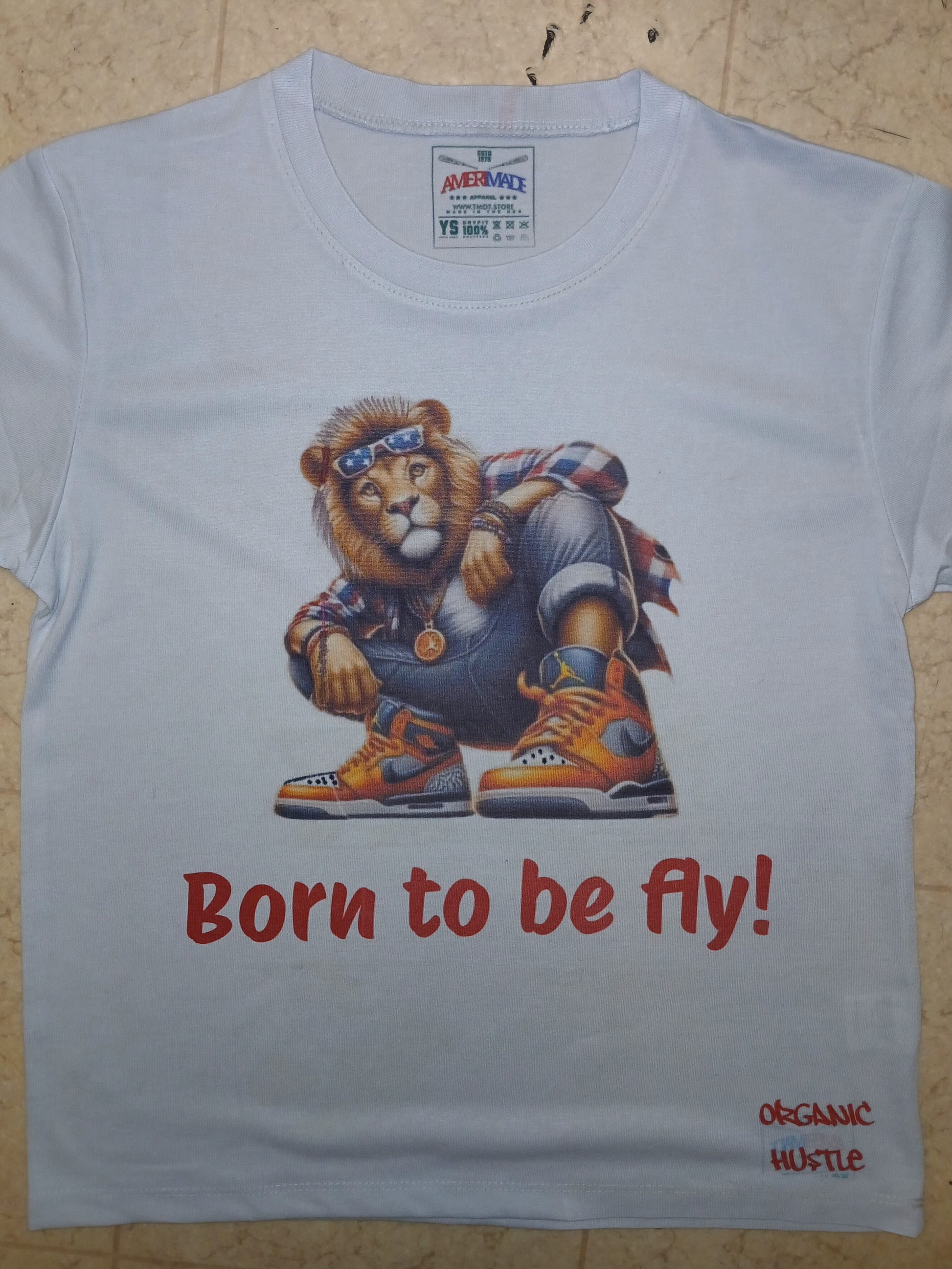 White t-shirt with a graphic of a lion wearing sneakers and a hat, with 'Born to be fly!' text, on a wooden surface.