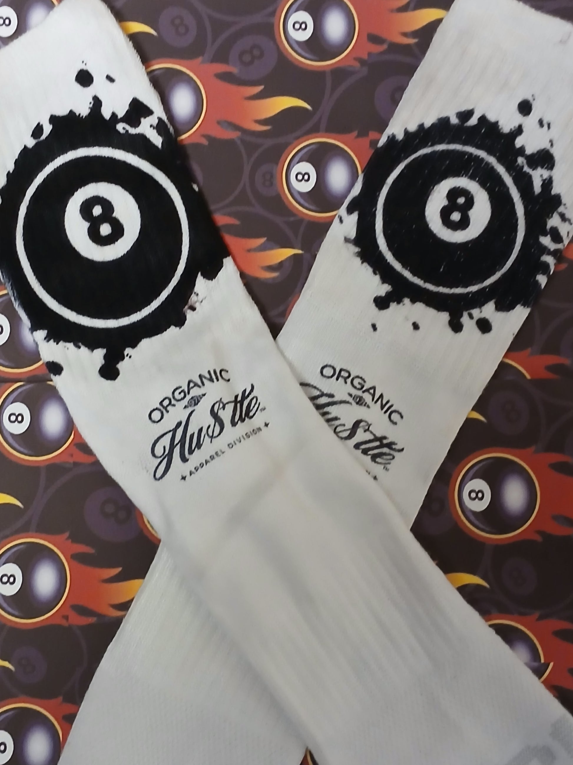 White socks with black eight-ball design and 'Organic Hustle' text on a patterned background.