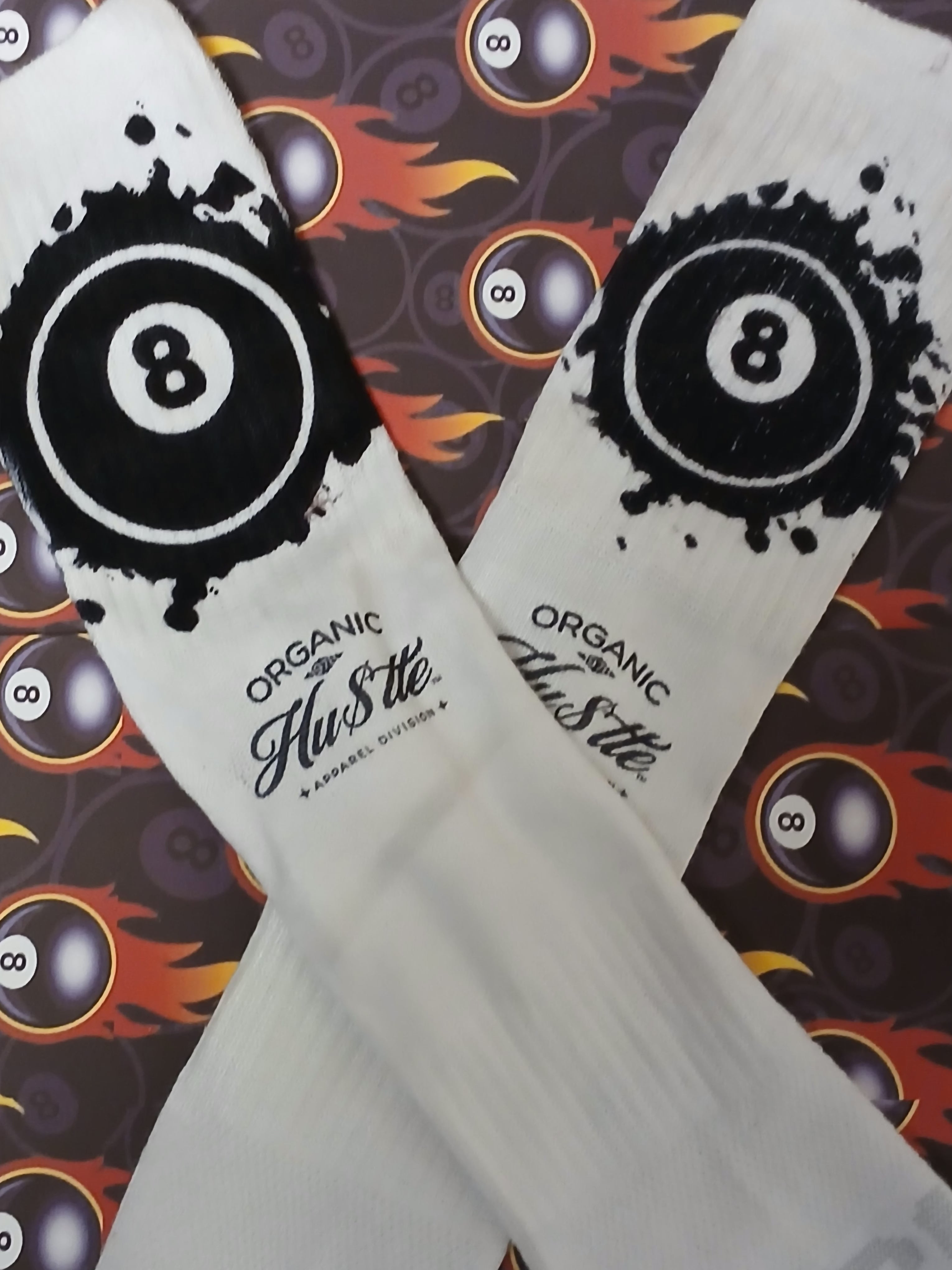 White socks with black eight-ball design and 'Organic Hustle' text on a patterned background.